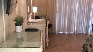 A room with a glass-top table, a potted plant, a lamp, a bed with a white headboard, and a curtain.