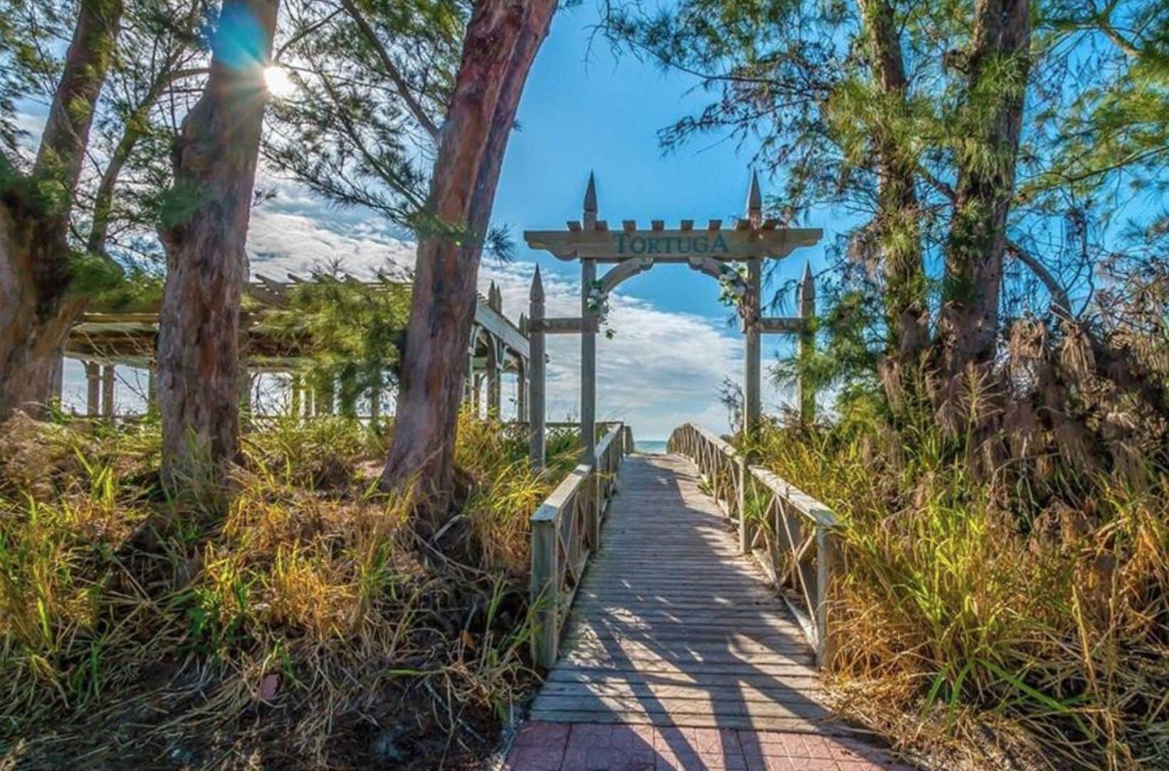 En træpromenade der fører til en pergola med ordet "FORTUGA" på, omgivet af træer og vegetation.