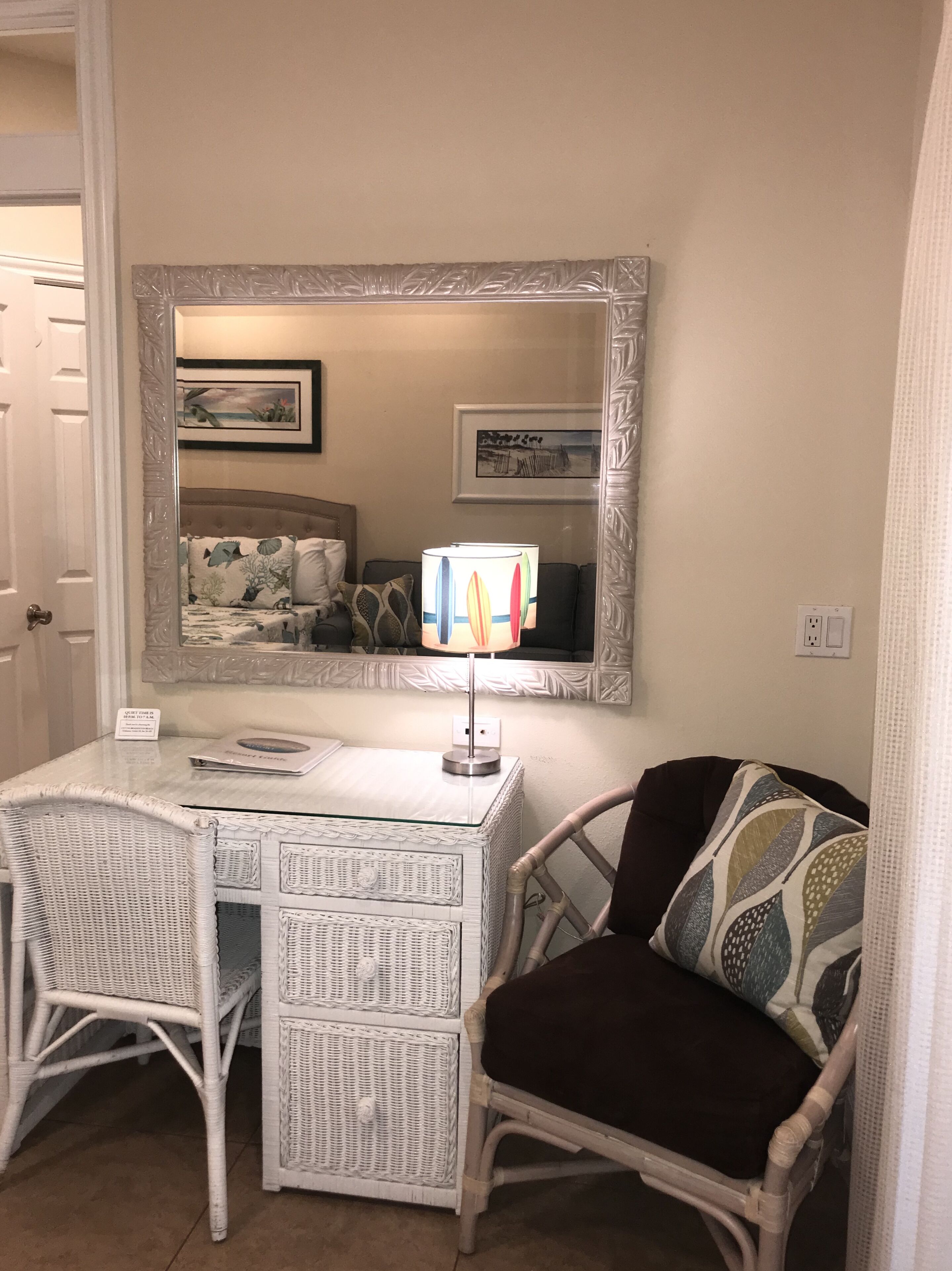 Una habitación con un escritorio de mimbre blanco, un espejo, una lámpara y una silla con cojín estampado.