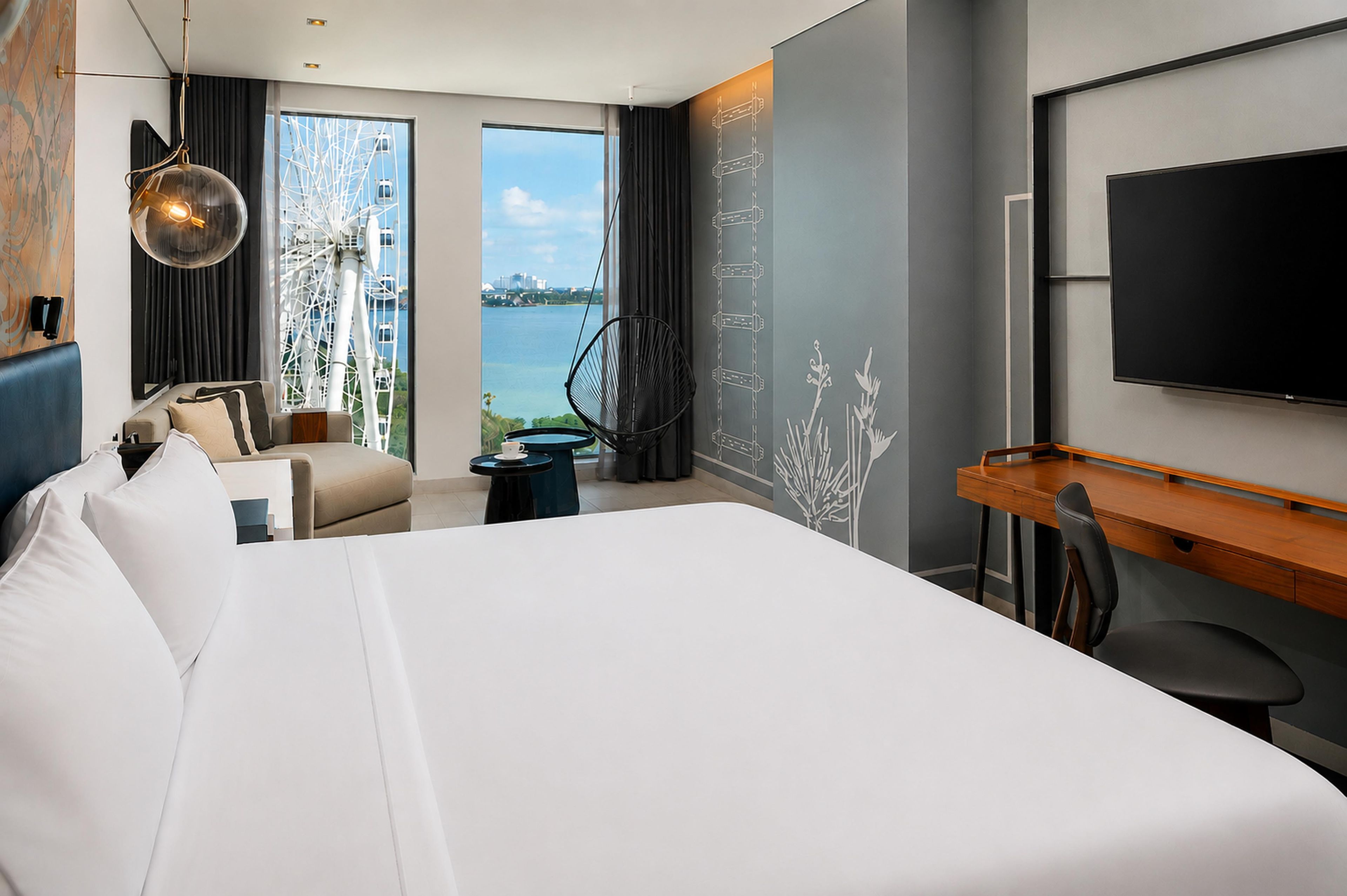 A hotel room with a large bed, a desk, a chair, a TV, and a view of a body of water and a Ferris wheel.
