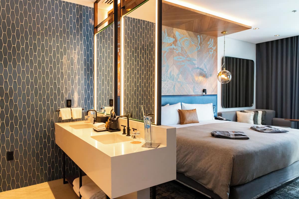 Una habitación de hotel moderna con una cama grande, un lavamanos doble y un espejo amplio.