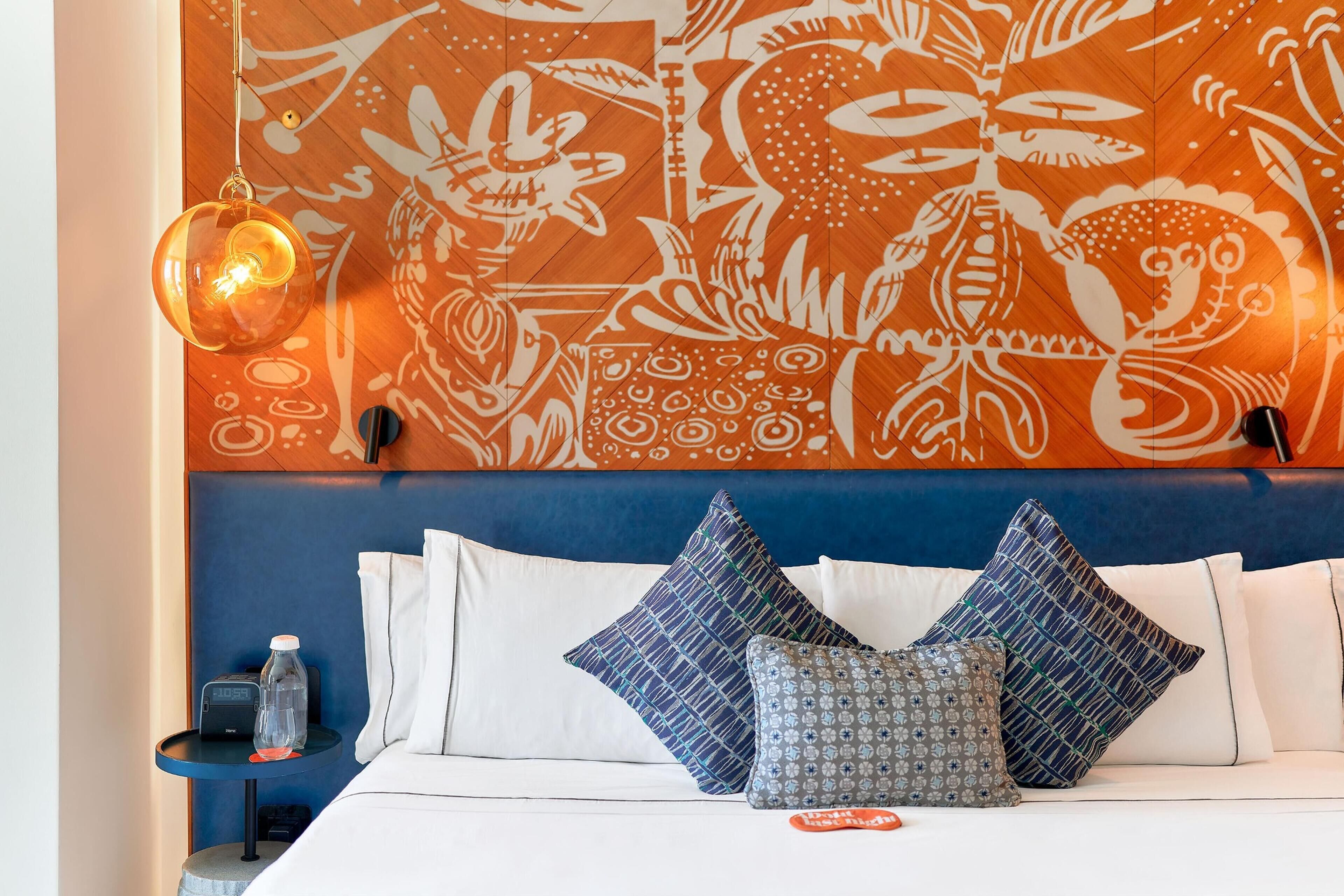 Habitación de hotel con cama, almohadas, un tapiz en la pared, una mesita de noche con una botella y un reloj, y una lámpara de pared.
