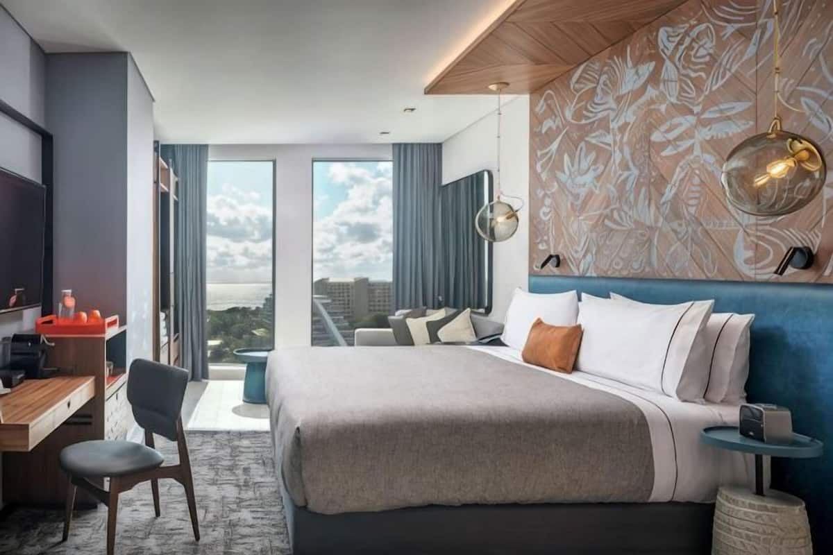 Habitación de hotel moderna con una cama grande, un escritorio, una silla, un televisor y vistas a la ciudad.