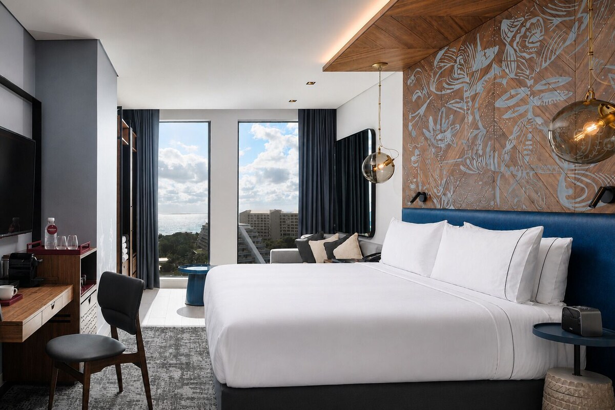 Habitación de hotel con una cama grande, un escritorio con silla, televisión y vistas a la ciudad a través de amplios ventanales.
