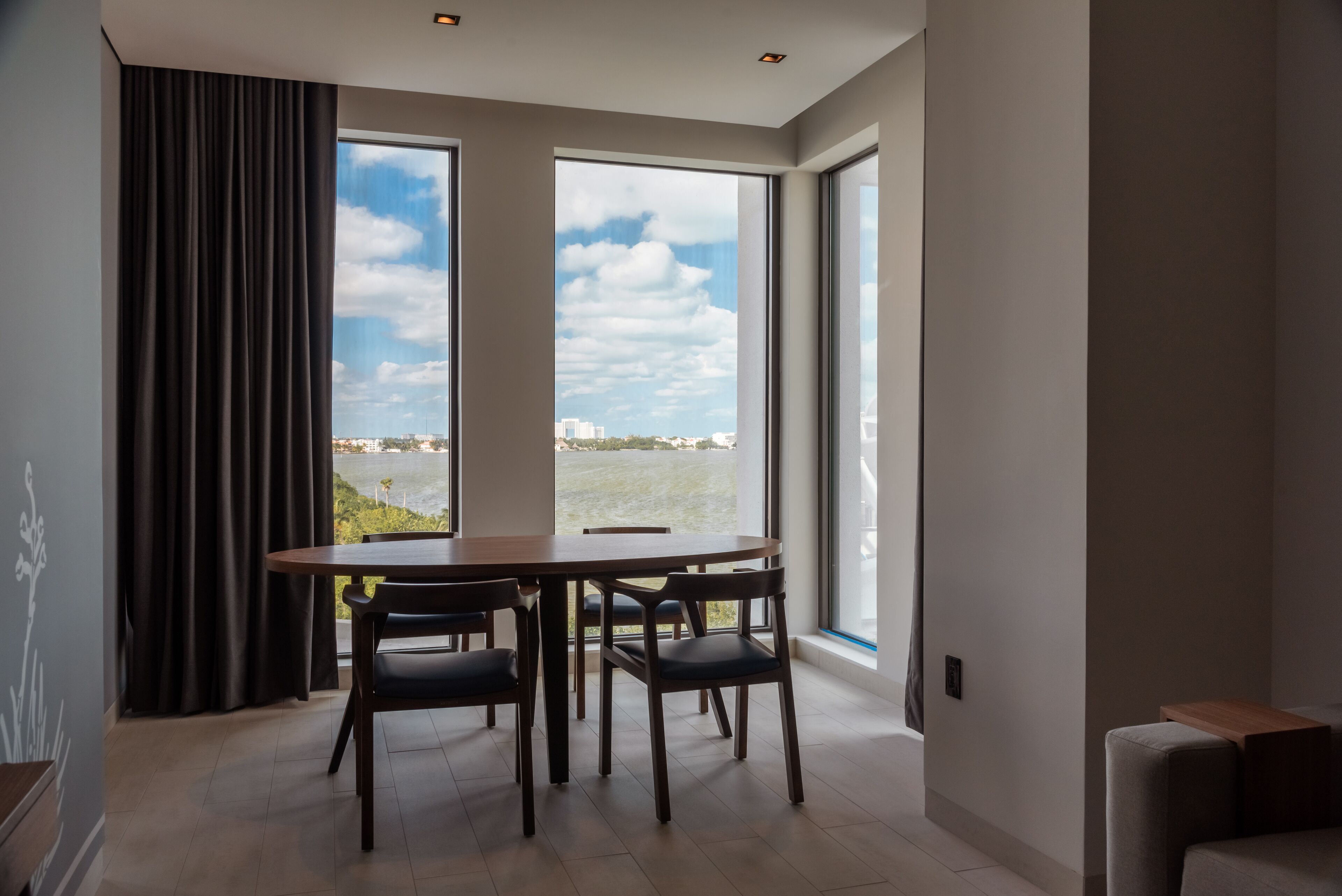 Un comedor moderno con una mesa y sillas, ventanas amplias con vistas a un cuerpo de agua y edificios, y un sofá gris en la esquina.