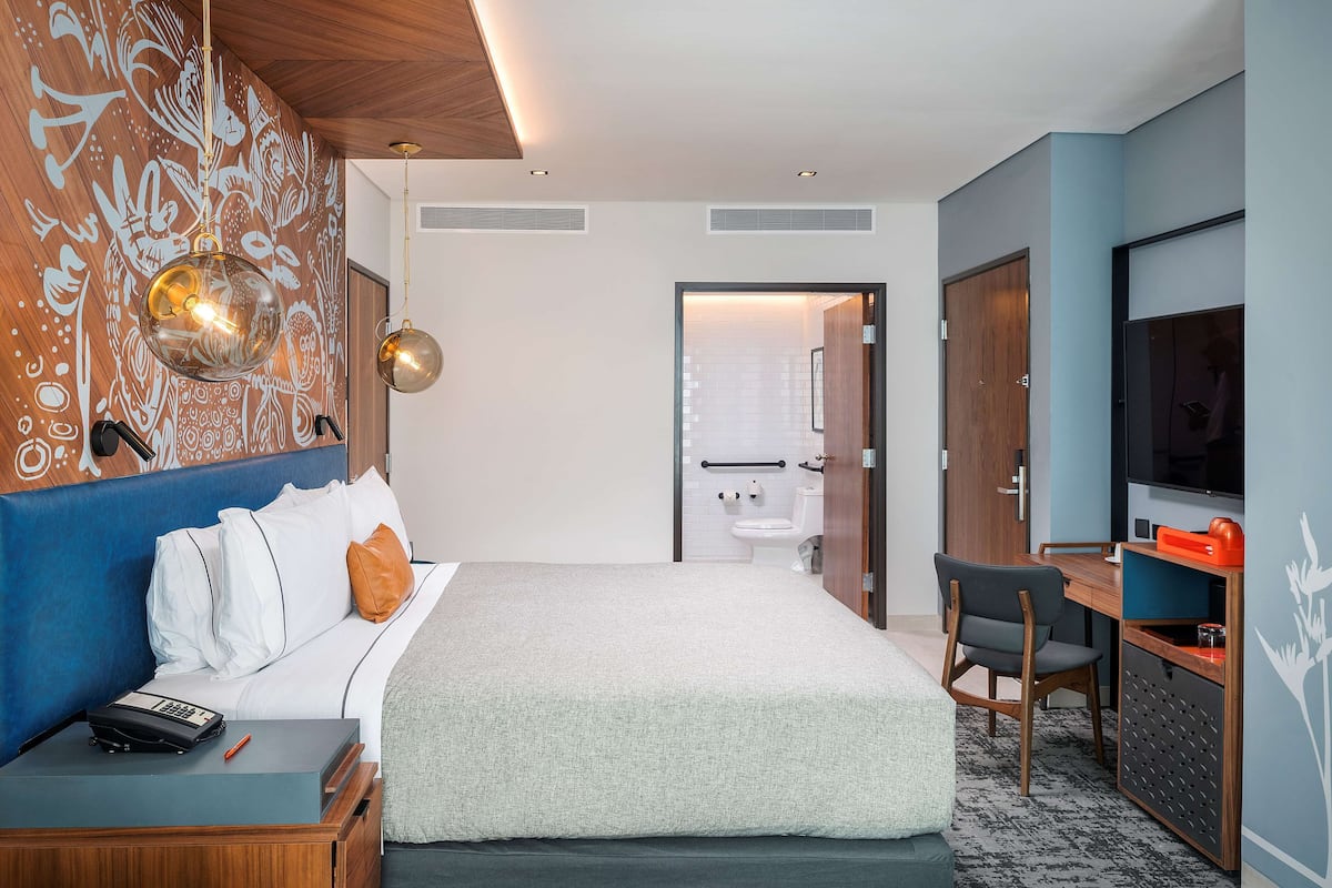 Habitación de hotel con cama, mesita de noche, escritorio, silla, televisión y baño visible a través de una puerta abierta.