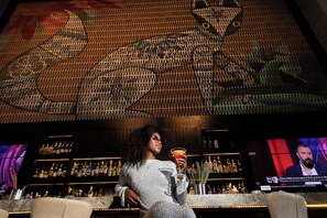 A bar area with a large screen displaying a news broadcast, shelves with bottles, and a seating area with a person holding a drink. - Canopy by Hilton Cancun La Isla (Cancun)
