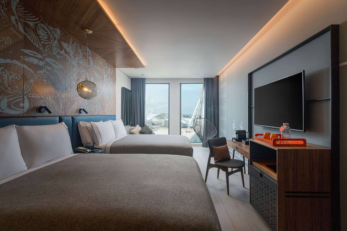 Habitación de hotel con dos camas, un escritorio con televisor y un amplio ventanal con vistas a la ciudad.