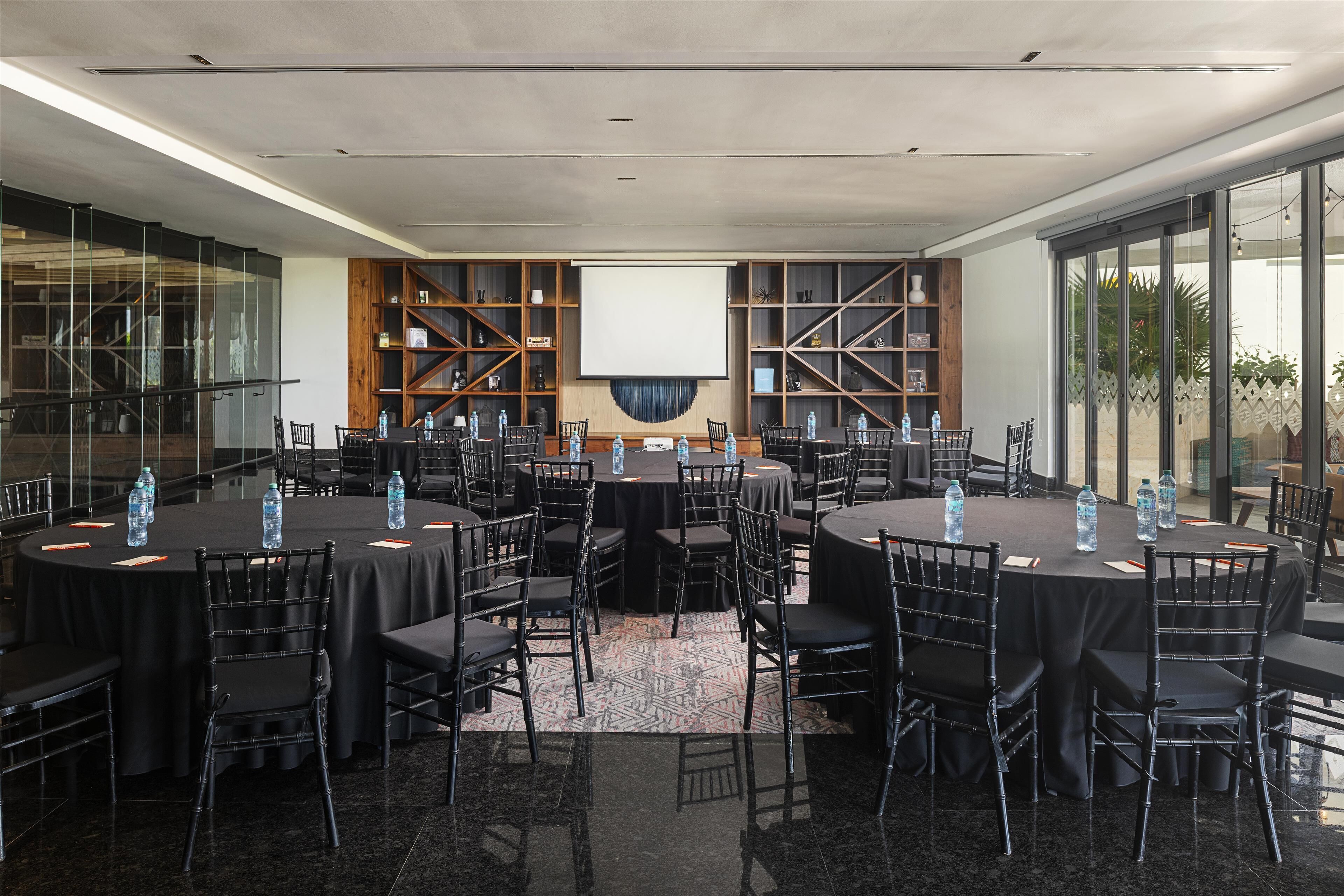 Un salón de conferencias con varias mesas y sillas, una pantalla grande y un muro de cristal con plantas.
