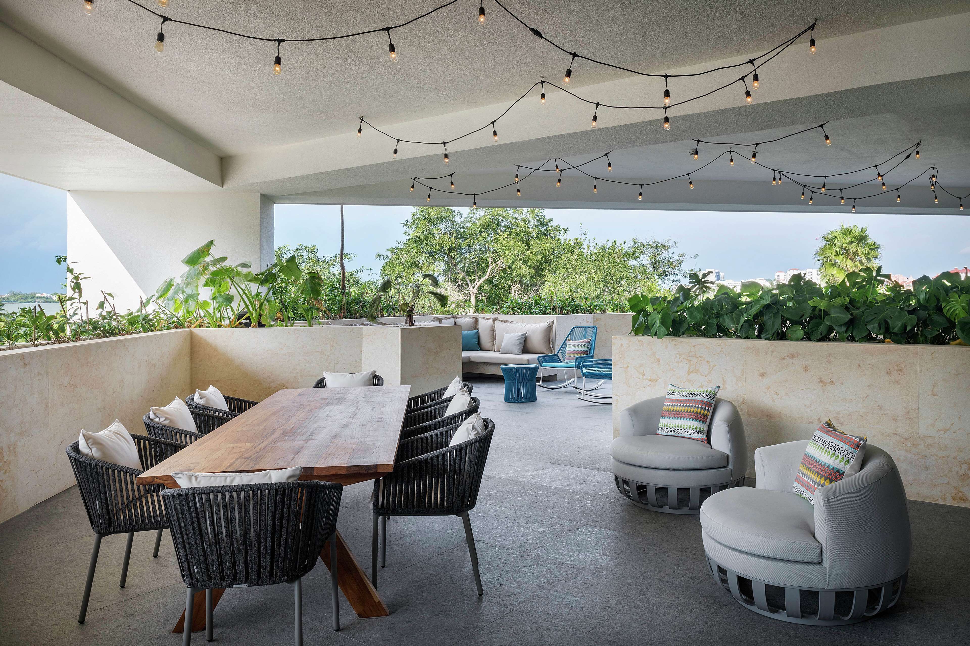 An outdoor patio with a wooden table and chairs, string lights, and a view of greenery and buildings.