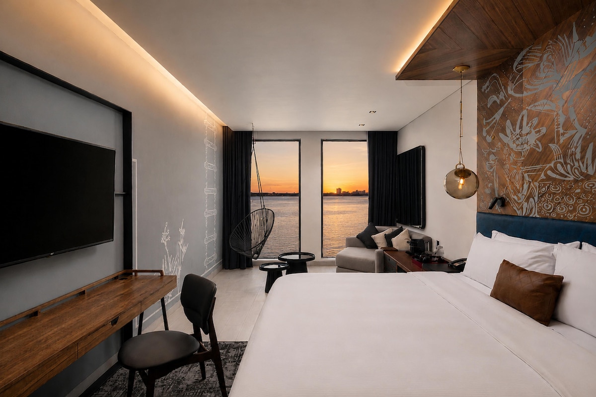 A modern hotel room with a large bed, a desk, a chair, a sofa, a TV, and a view of the sunset over the water.