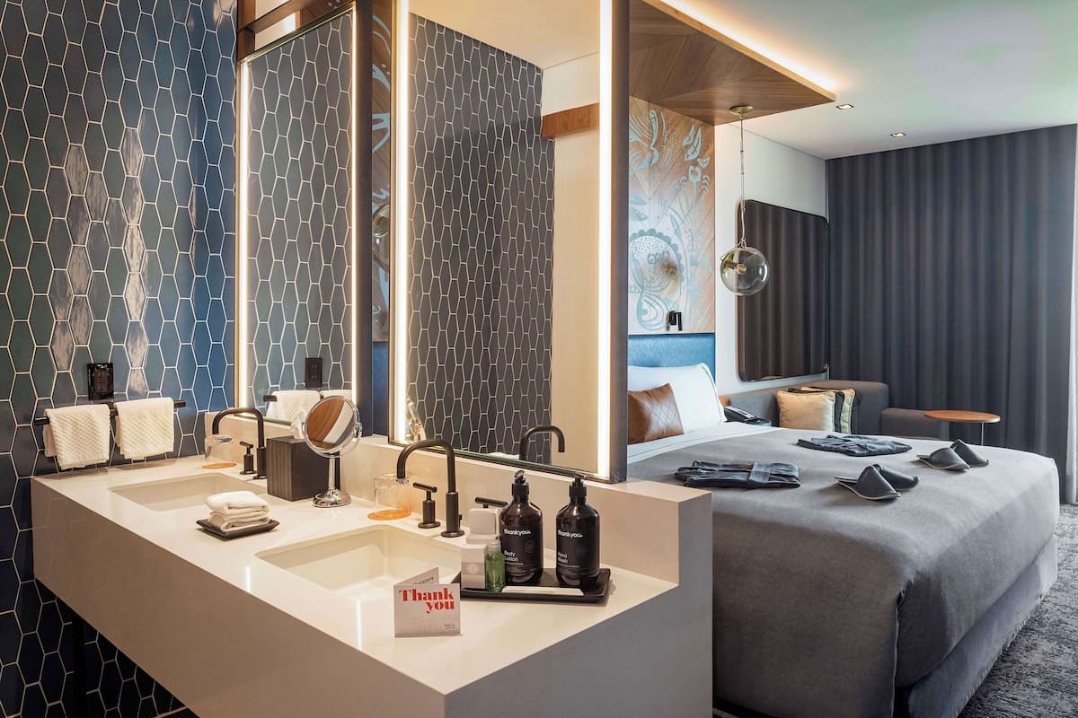 Una habitación de hotel moderna con una cama grande, un baño con lavamanos doble y un espejo amplio.