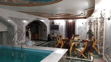 A luxurious indoor swimming pool area with marble flooring, a pool with clear blue water, wooden chairs, and a ceiling mural.