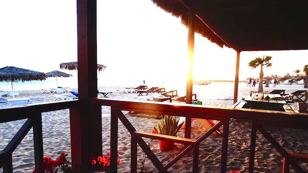Une vue sur la plage depuis une terrasse en bois donnant sur le sable et l’océan, avec des chaises de plage et des parasols visibles.