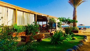 Un bâtiment situé en bord de mer, doté d’un espace extérieur abrité avec des sièges, entouré d’une végétation luxuriante et d’un palmier.