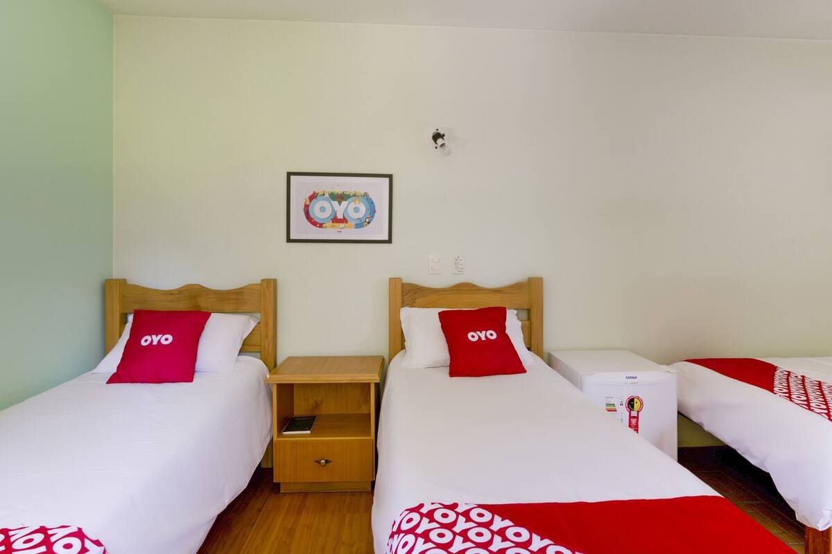 Ein Hotelzimmer mit zwei Einzelbetten, jeweils mit einem hölzernen Kopfteil und einem roten Kissen mit OYO-Branding. Zwischen den Betten steht ein kleiner Nachttisch mit einem Buch darauf. Über den Betten hängt ein gerahmtes Bild mit dem OYO-Logo an der Wand.