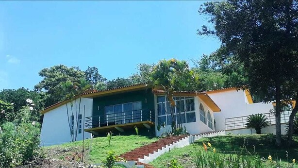 A two-story house with a staircase leading to the entrance, surrounded by lush greenery and trees.