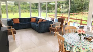 A modern living room with a sectional sofa, wooden chairs, a dining table with a patterned tablecloth, and large windows offering a view of a garden.