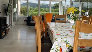 A dining area with a table set for four, large windows offering a view of greenery and hills, and a chandelier hanging from the ceiling.