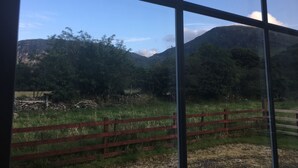 A view through a window with a metal frame, featuring a red wooden fence, green grass, and a mountainous landscape in the background.