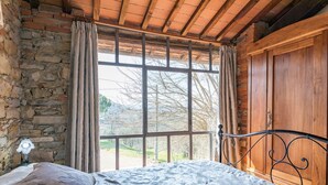Ein Schlafzimmer mit Steinwand, Holzboden, einem großen Fenster mit Vorhängen, einem Metallbettgestell und einem Holzschrank.