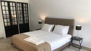 Una cama bien tendida con cubrecama color beige y almohadas blancas, flanqueada por dos mesitas de noche con lámparas, en una habitación con suelo de baldosas y un ventanal con paneles de vidrio esmerilado.