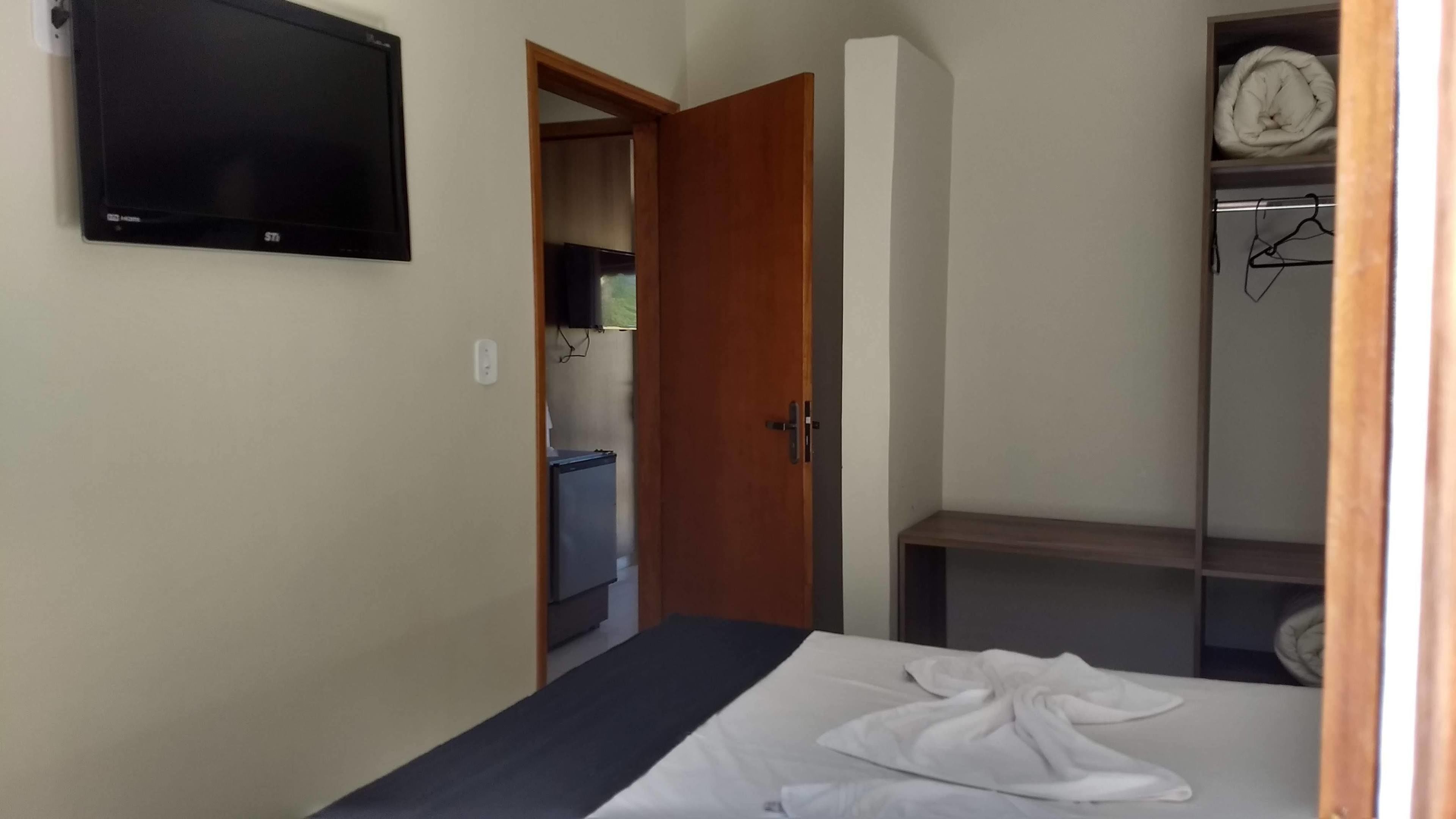 Quarto com cama, televisão fixada na parede, guarda-roupa e porta que dá para outro espaço.