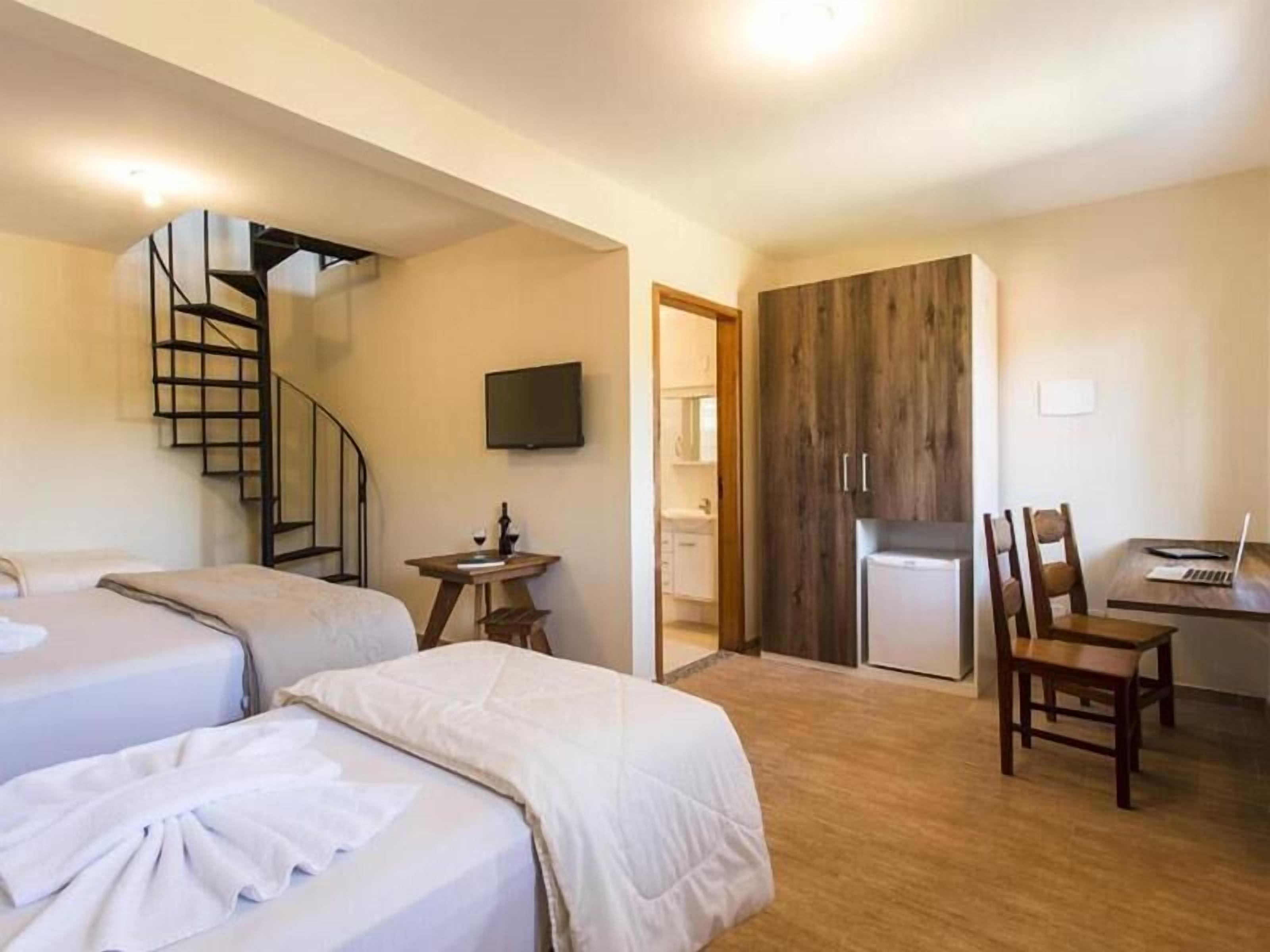 Quarto de hotel com duas camas, uma escrivaninha com um laptop, uma televisão, um guarda-roupa de madeira e uma escadaria em espiral.