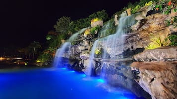 Uma cascata noturna com água caindo sobre pedras, rodeada por vegetação exuberante.