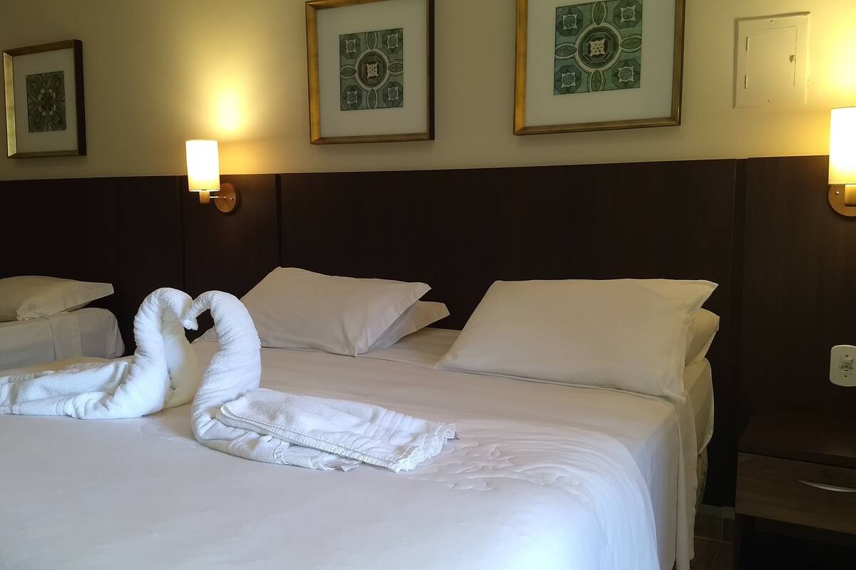A hotel room with two beds, each with white linens and pillows. There are wall-mounted lamps on either side of the beds.