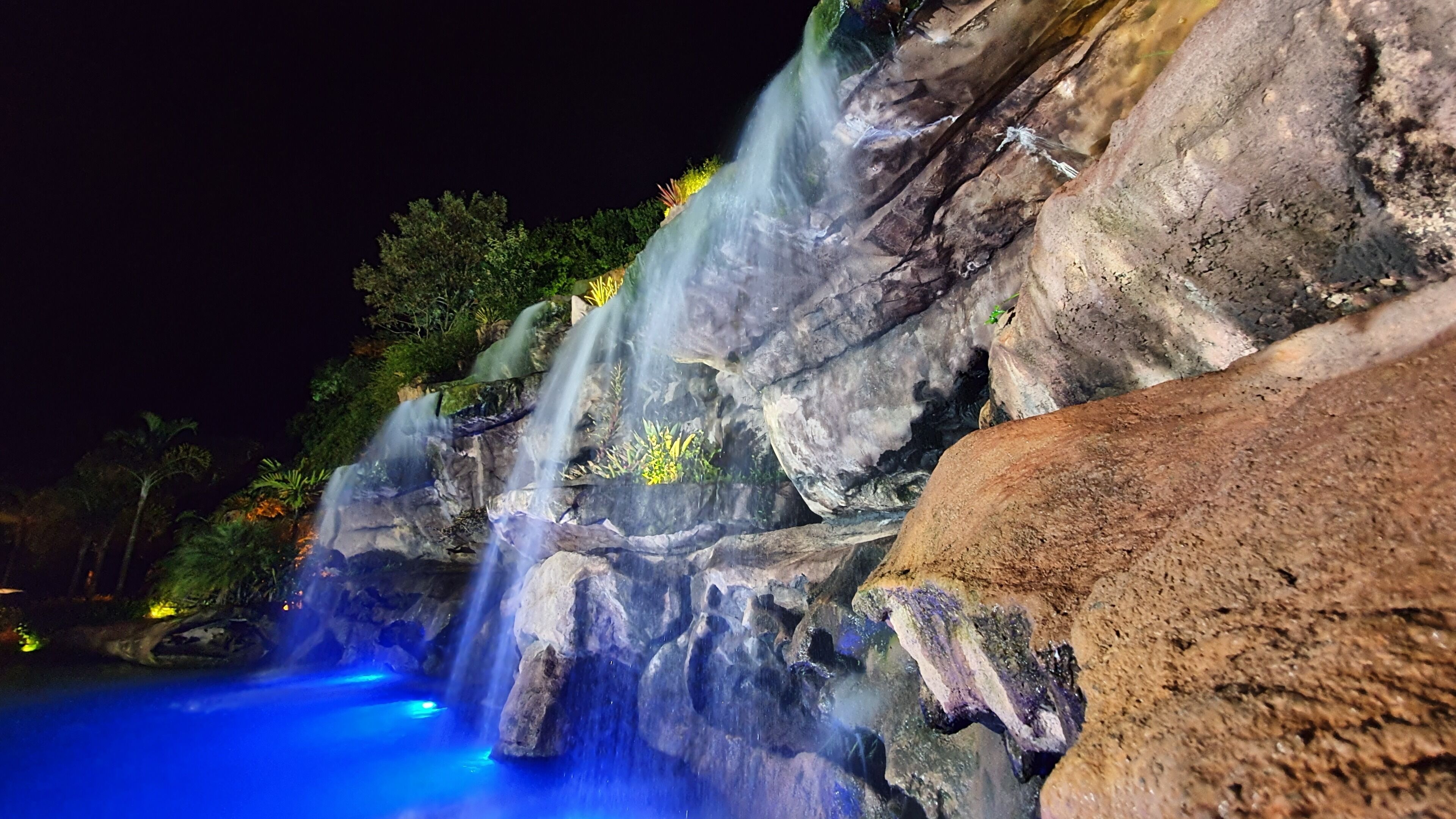 Una cascada nocturna con plantas iluminadas y una piscina en la base.