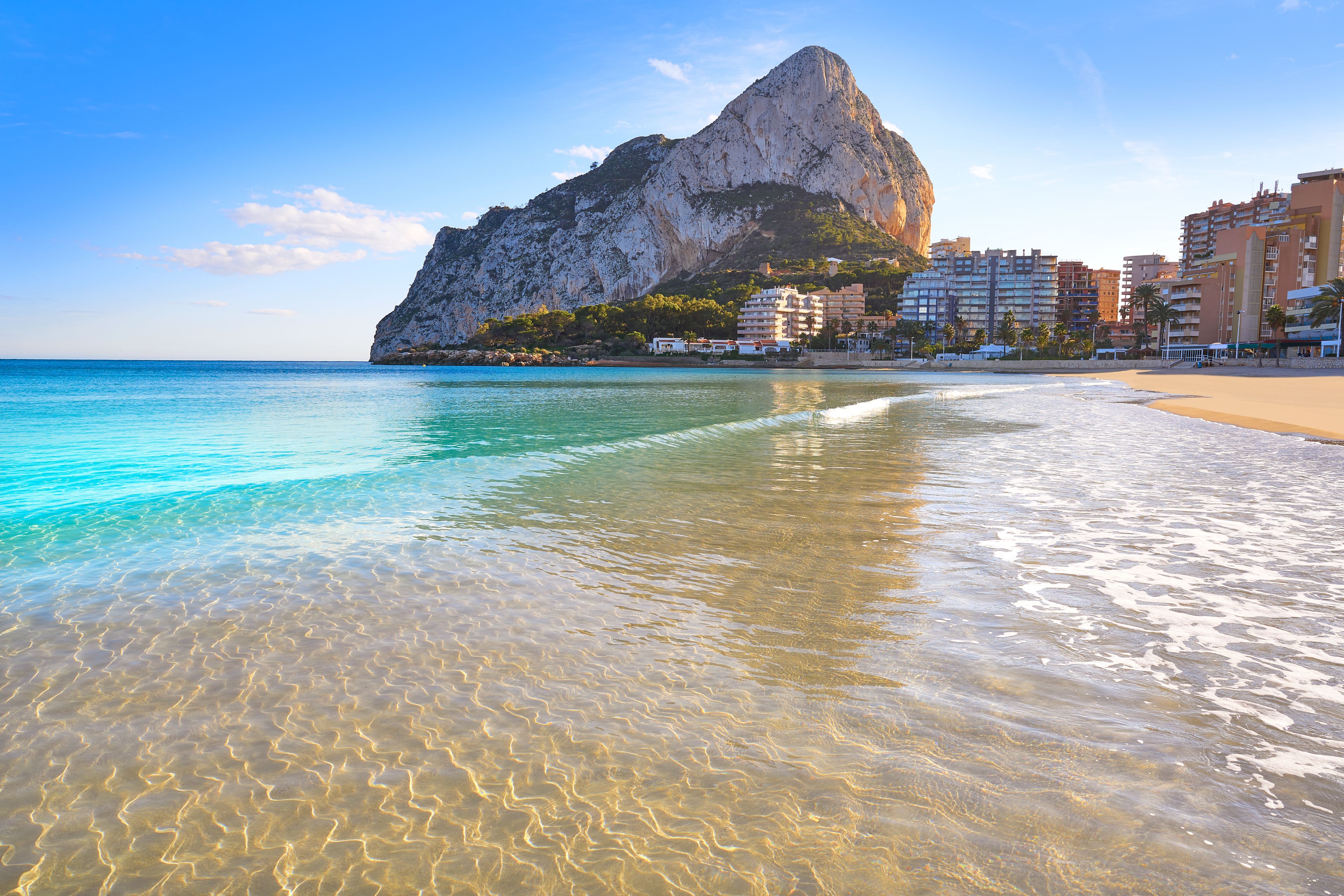 A beach with clear turquoise water, a prominent rock formation, and buildings in the background.
