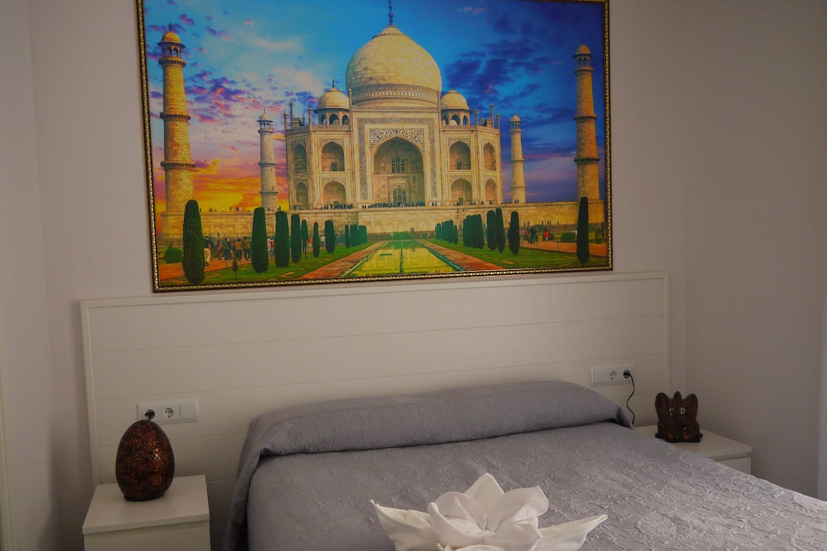 A hotel room with a large painting of the Taj Mahal on the wall, a neatly made bed with a white flower arrangement, and a vase on the nightstand.