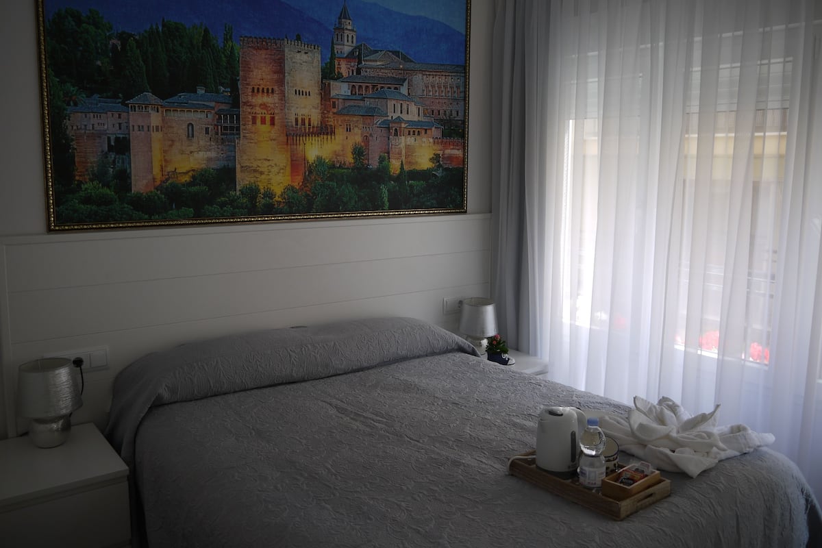 A hotel room with a large painting of a castle, a bed with a gray blanket, a bedside table with a lamp, and a tray with toiletries.