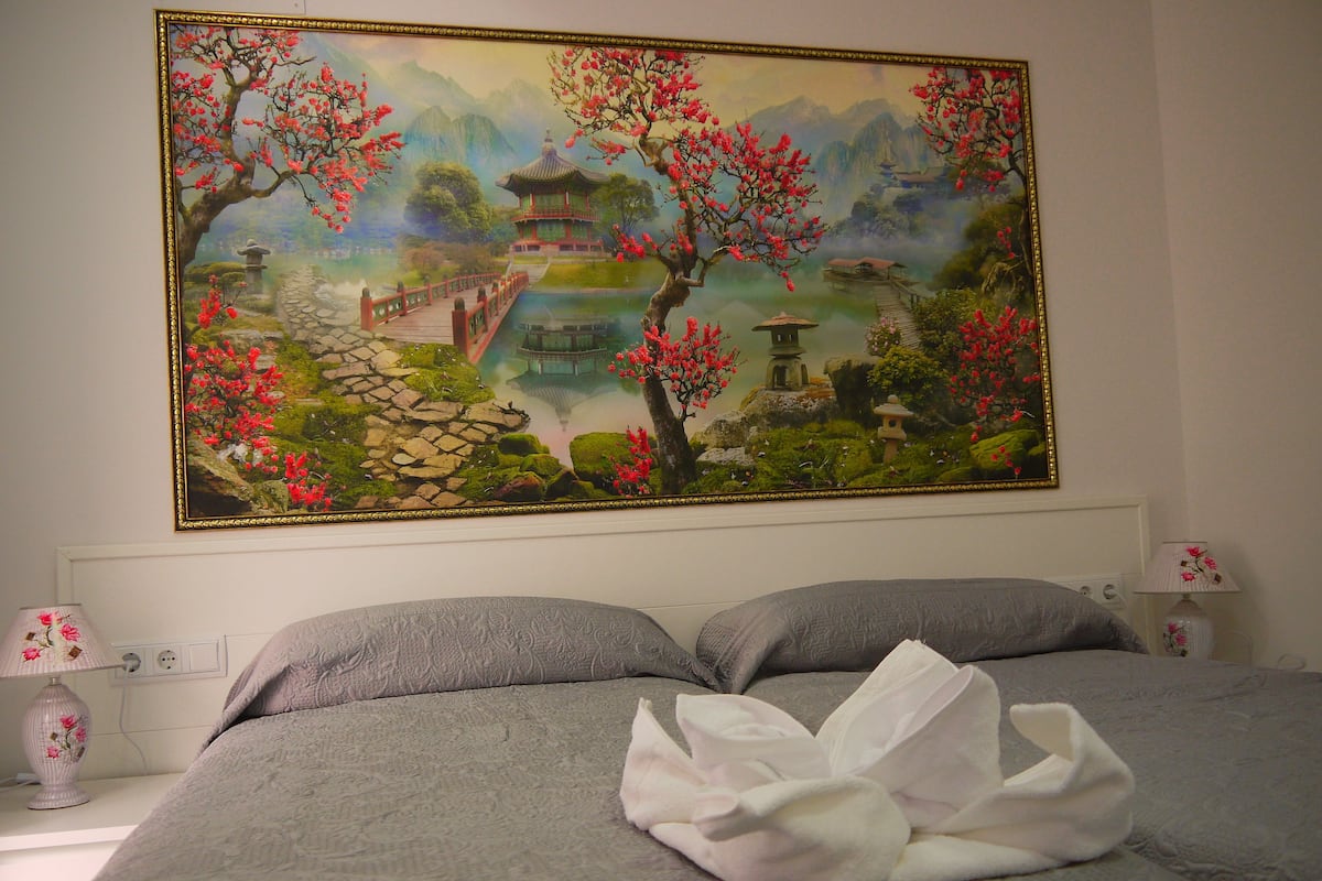 A bedroom with a large painting of a traditional landscape, a bed with a white towel, and a nightstand with a lamp.