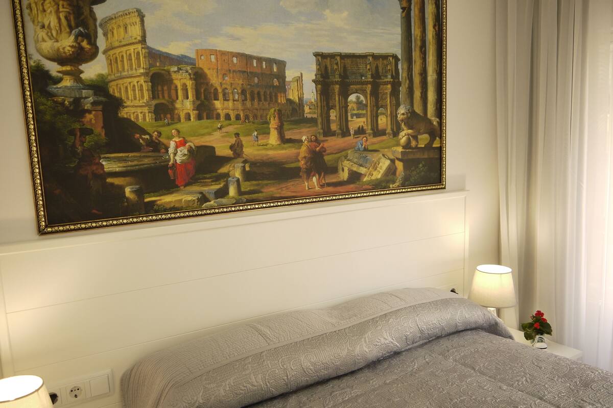 A hotel room with a large painting of the Colosseum, a bed with a grey blanket, two bedside lamps, and a small vase with flowers on the nightstand.