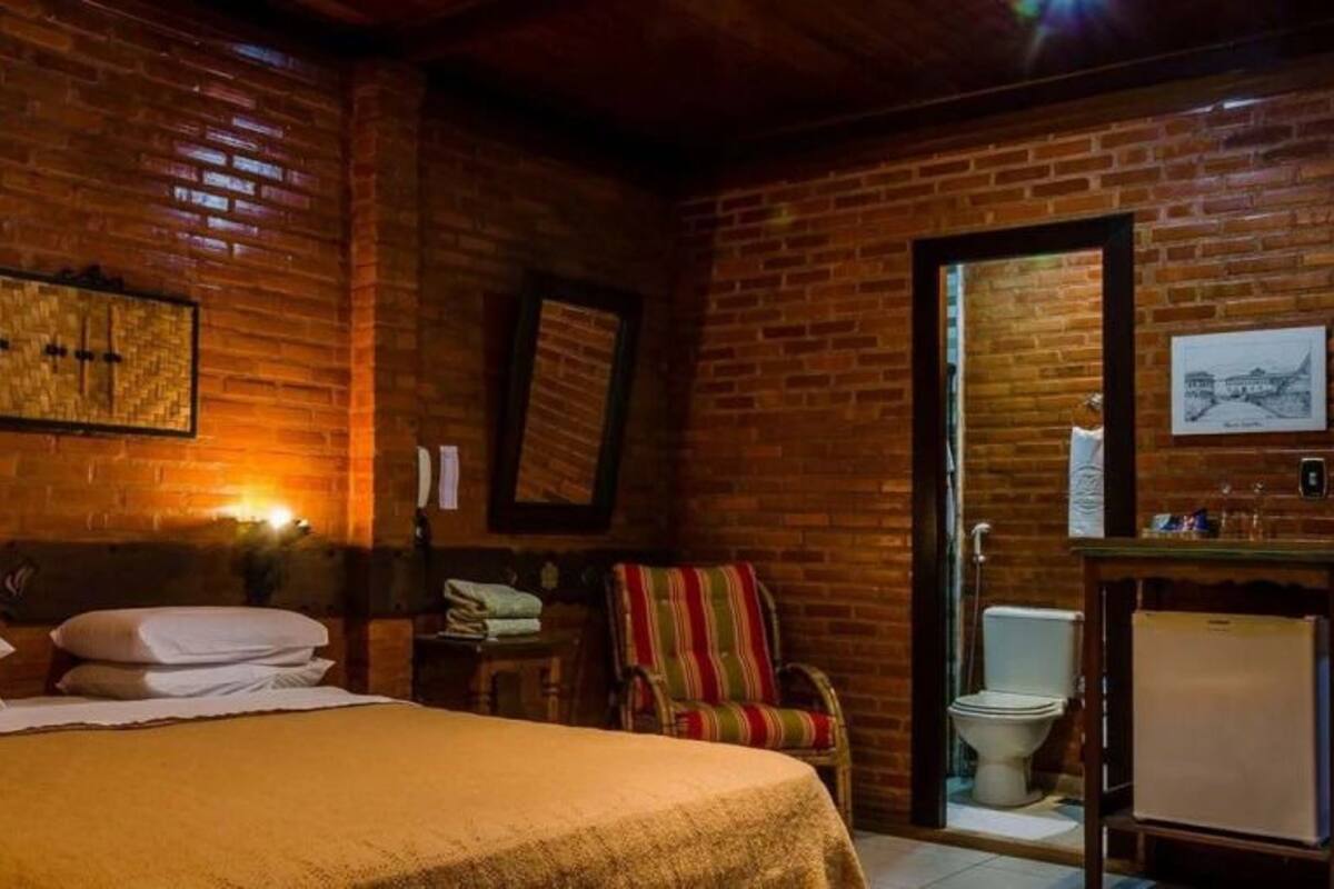 A cozy room with a brick wall, a bed with white linens, a striped chair, a small table, and a toilet.