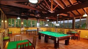 A pool hall with a green pool table, wooden tables, and chairs. There are windows with views of greenery outside.