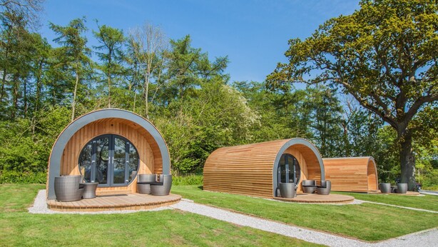 Twee houten cabin-achtige accommodaties met grote ramen en zitplaatsen, gelegen in een verzorgd gazon met bomen.