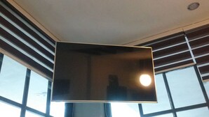 Un téléviseur à écran plat fixé au plafond dans une pièce dotée de grandes fenêtres et de stores.