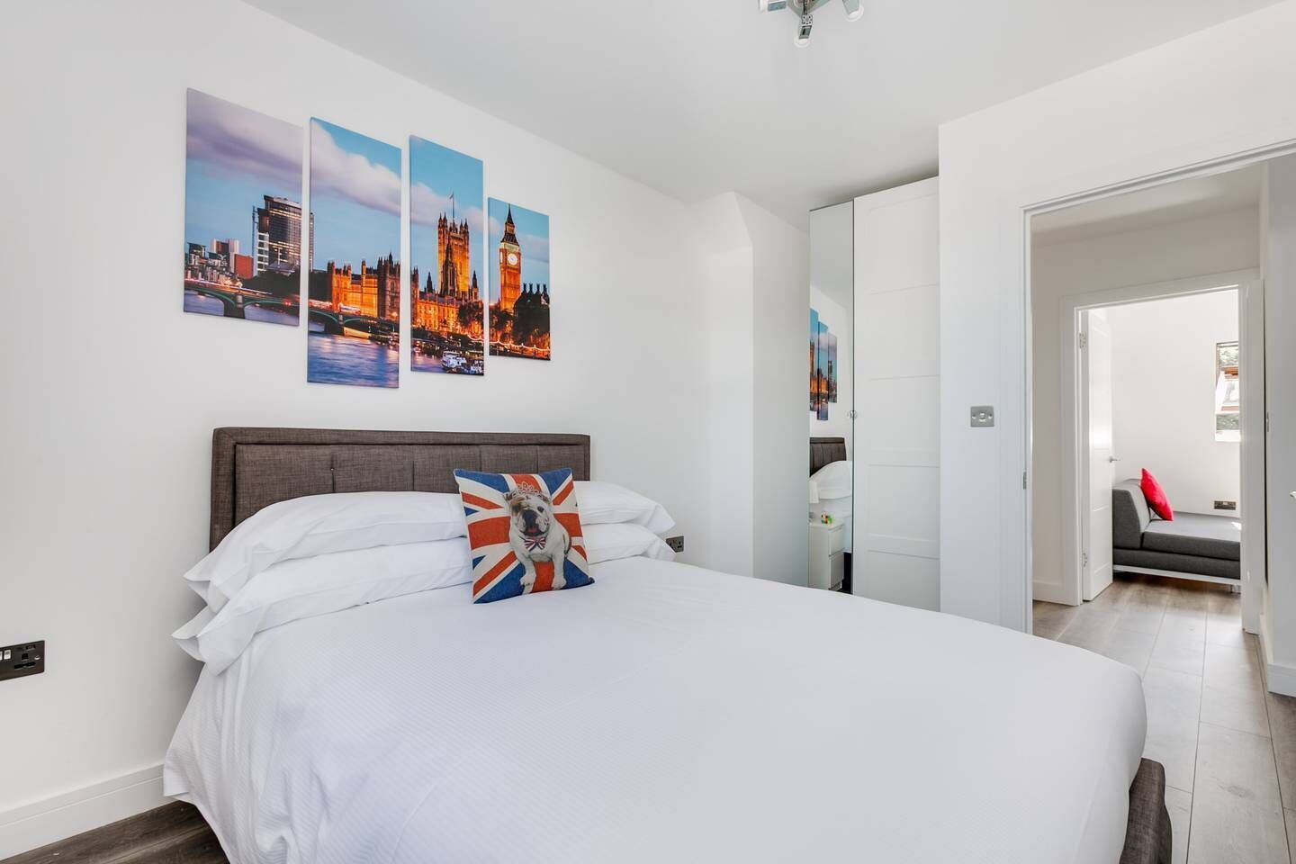 Una habitación de hotel moderna con una cama grande, ropa de cama blanca, una almohada con la bandera Union Jack y tres cuadros de paisajes urbanos en la pared.
