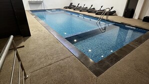 Una piscina interior con superficie de agua cristalina, rodeada de suelo y paredes con azulejos. Hay sillas de descanso y una escalera que da a la piscina.