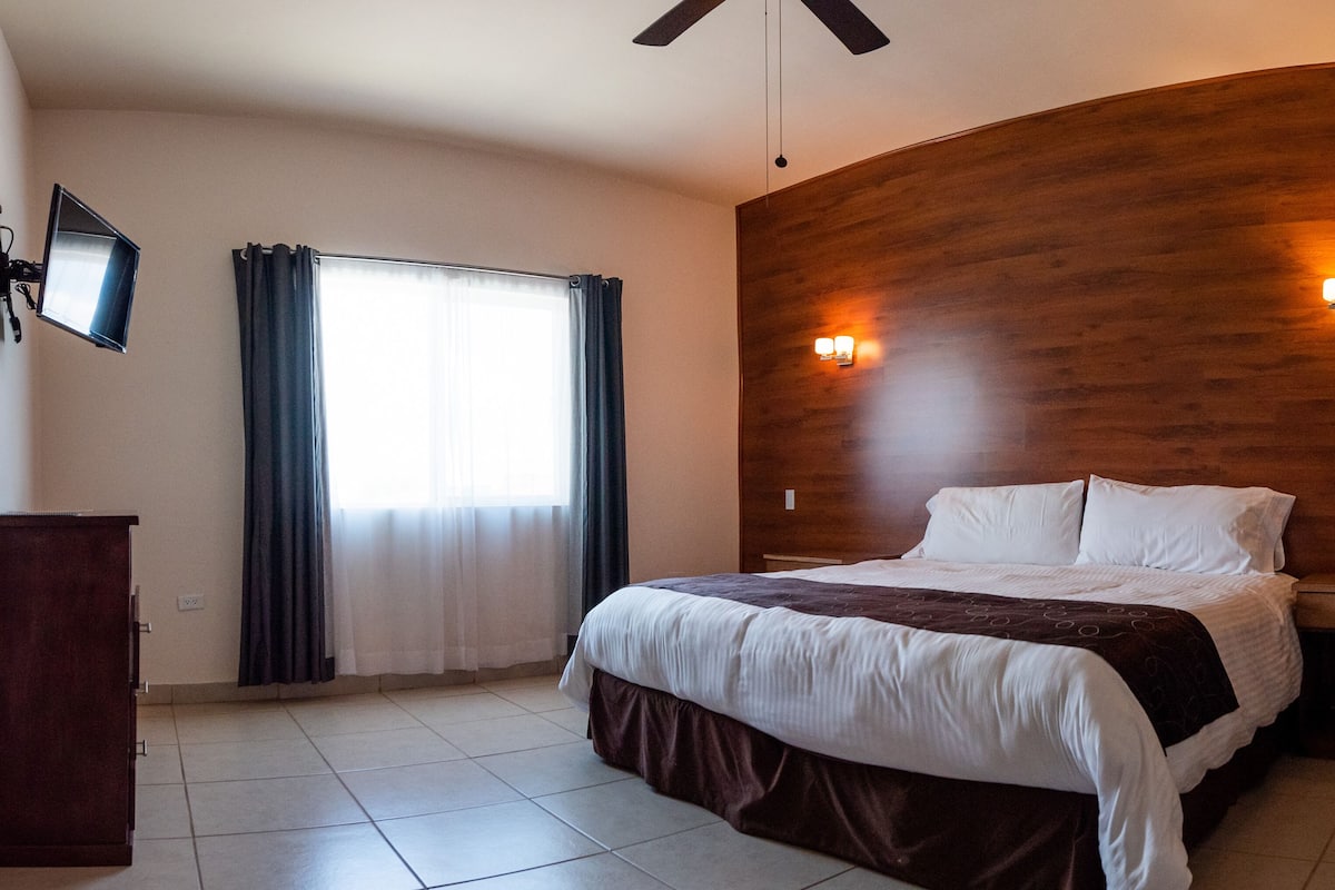 A hotel room with a bed, a nightstand, a desk, a ceiling fan, a wall-mounted TV, and a window with curtains.