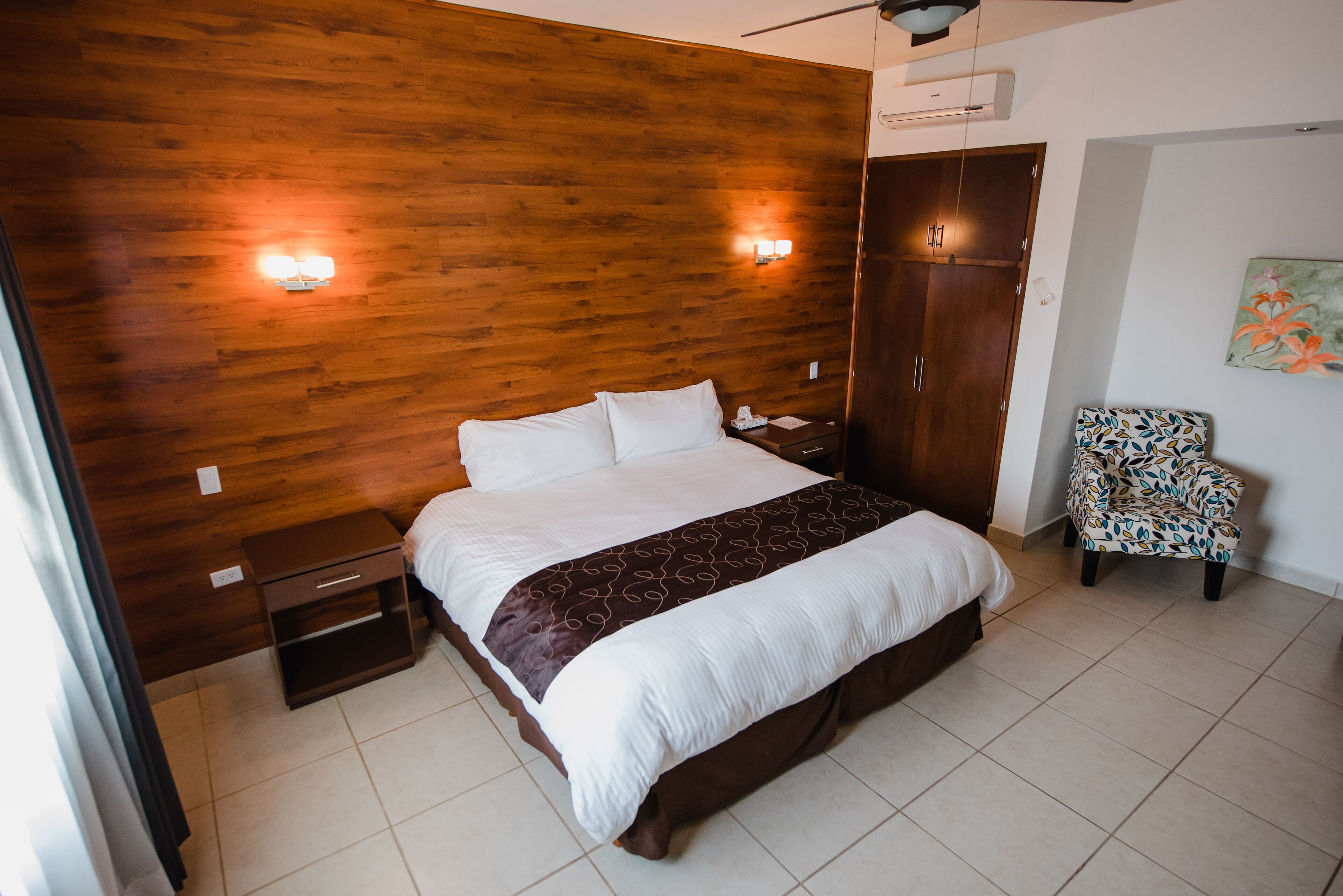 Une chambre d’hôtel avec une tête de lit en bois, un grand lit avec une literie blanche, une table de chevet, une chaise et une armoire.