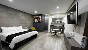 Una habitación de hotel moderna con una cama grande, un escritorio con dos sillas, una kitchenette y un televisor montado en la pared.