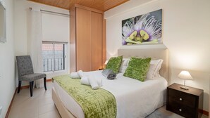 A bedroom with a bed, a chair, a nightstand, a painting, and a window with a view.