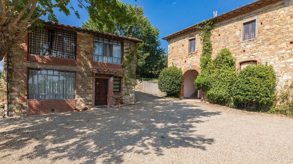 A stone building with a gravel driveway, a wooden door, and a window with a checkered curtain.
