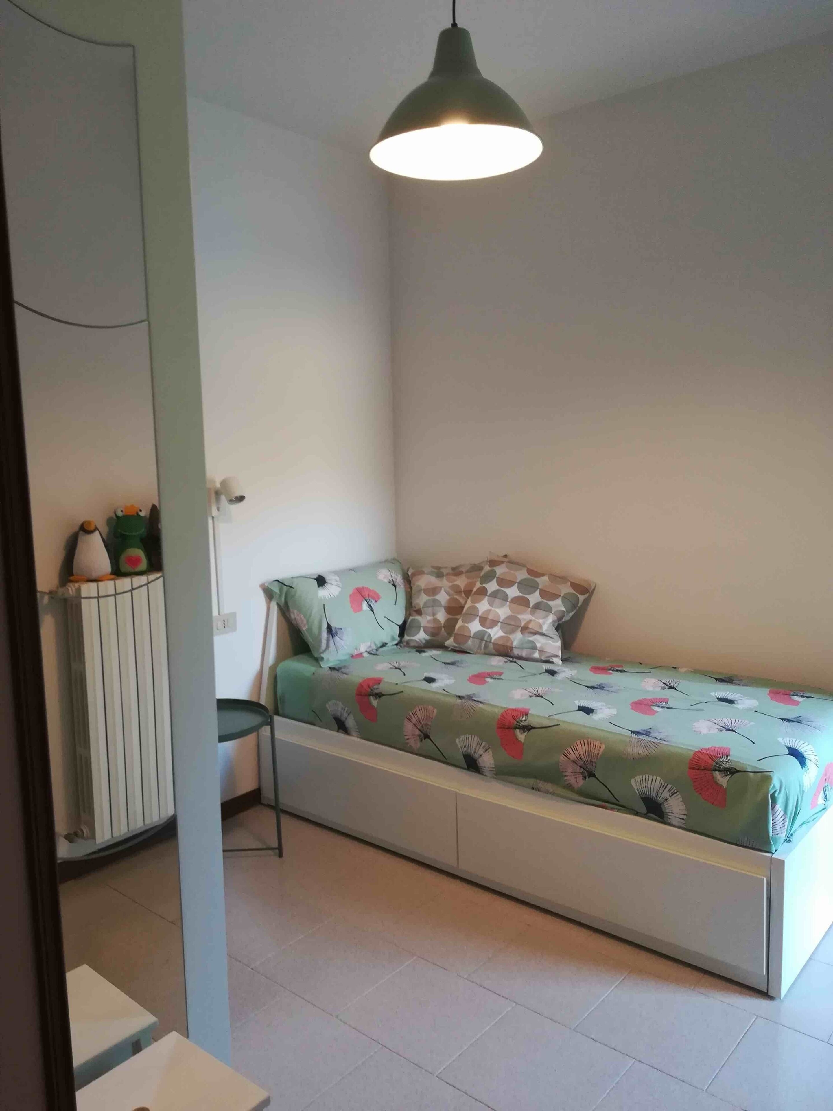A small, single-bed room with a green floral bedspread, a hanging pendant light, and a white radiator.