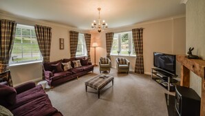 A living room with a sofa, armchairs, a coffee table, a TV, and curtains. - MacKenzie Cottage - Family Friendly Cottage on the Beautiful Blairquhan Estate (Maybole)