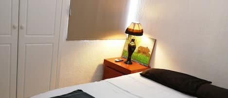 En snyggt bäddad säng med ett svartvitt överkast, ett litet sängbord med en lampa, en fotoram och en luftkonditioneringsenhet som är monterad på väggen.